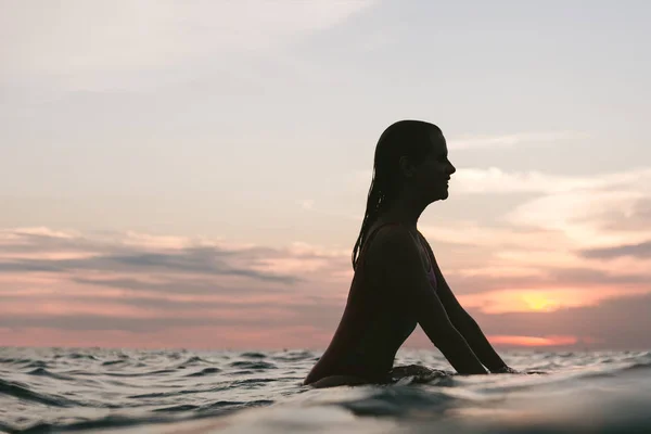 Vista lateral da silhueta de mulher descansando sobre prancha de surf no oceano ao pôr do sol — Fotografia de Stock