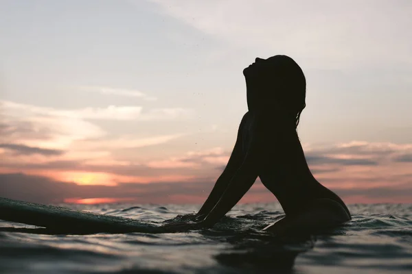 Silueta de mujer descansando sobre tabla de surf en el océano al atardecer - foto de stock