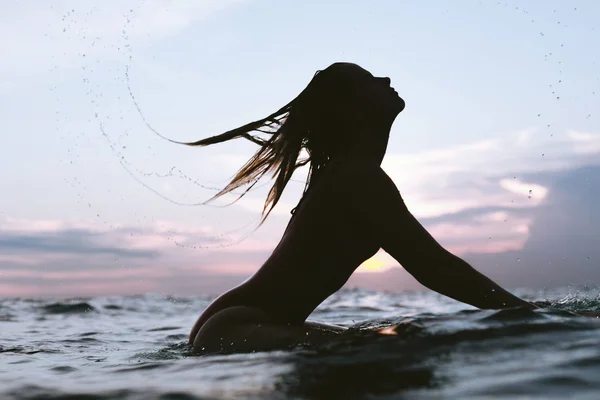 Silhouette di donna frustate capelli mentre si riposa su tavola da surf in mare al tramonto — Foto stock