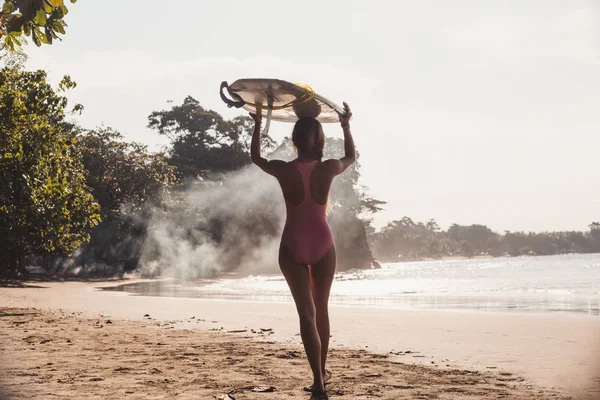 Вид сзади женщины с доской для серфинга на голове во время прогулки по береговой линии — стоковое фото
