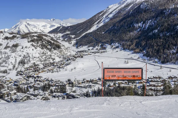 Signpost written in various languages warning of expert ski run ahead. View of Thuile town in valley below Valle d'Aosta, Italy — Stock Photo, Image