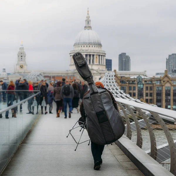 Rücken eines nicht identifizierbaren Straßenmusikers, der ein umgedrehtes Cello auf der Millenium Bridge mit Blick auf die St. Paul Kathedrale in London, England, Großbritannien trägt. — Stockfoto