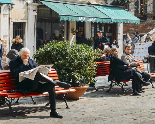 Un hombre mayor lee un periódico italiano local en un banco cuadrado rodeado de conmoción — Foto de Stock