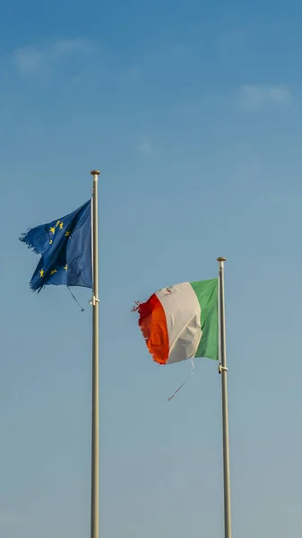 Italy flag and Europe flag waving together on a mast in isolated the blue sky background. Concept for financial treated, unique currency and financial bond despite the torn sides.