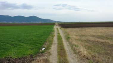 Arka planda tepeler olan tarımsal tarlalar arasında toprak bir yol üzerinde alçak uçuş