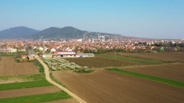 Banliyöleri ve çevresindeki tarlalarıyla bir tepenin altındaki bir şehrin panoraması - insansız hava aracı görüntüsü ilerliyor