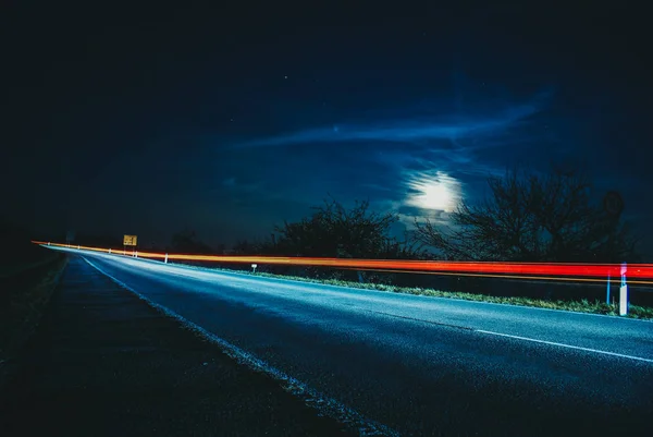 red lights of a fast approaching car in a street on the countryside in a blue dark sky night with the moon out and trees in the background