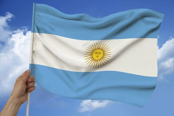 Mano masculina sostiene sobre el fondo del cielo con nubes la bandera nacional de Argentina sobre una lujosa textura de satén, seda con olas, primer plano, espacio de copia, concepto de viaje, economía, política —  Fotos de Stock