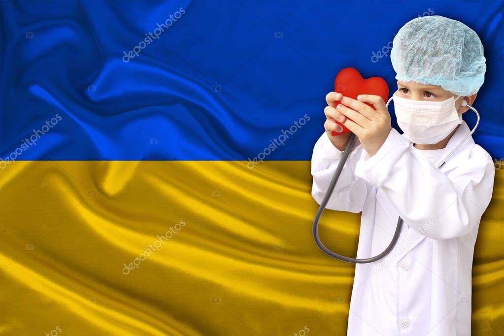 child, boy, in a white doctor’s coat, hat and mask attached a stethoscope to a red heart model, background flag Ukraine close-up, face focus, medical concept, cardiology