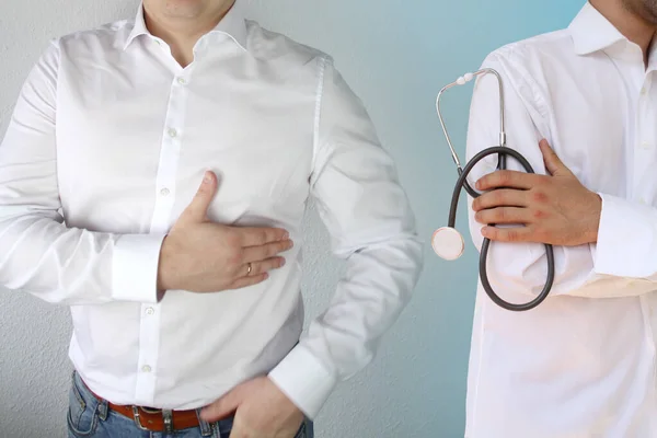 Man in a white shirt holds on to his chest in the heart area, next is a doctor in a white coat with a stethoscope, concept of health problems, illness, first aid — Stock Photo, Image