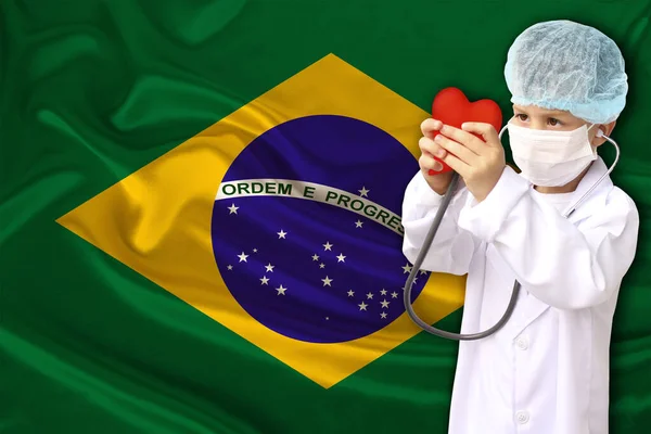 Child, boy, in a white doctor’s coat, hat and mask attached a stethoscope to a red heart model, Brazil flag background, close-up, face focus, medical concept, cardiology, copy space — Φωτογραφία Αρχείου