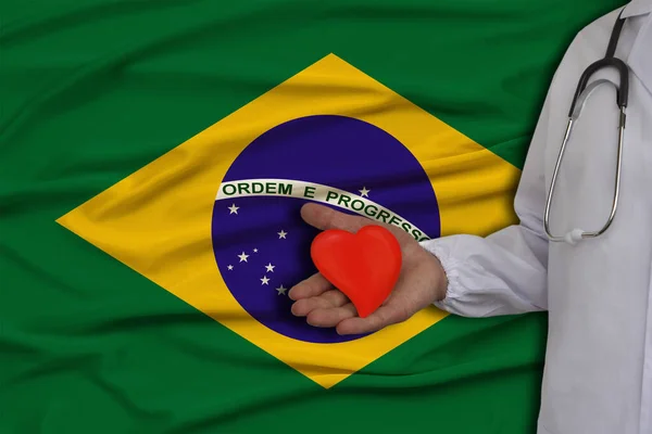 Φωτογραφία ενός γιατρού με στηθοσκόπιο με ένα μοντέλο της καρδιάς στο χέρι σχετικά με το υπόβαθρο της εθνικής σημαίας της Βραζιλίας, έννοια της υγειονομικής περίθαλψης, καρδιολογία, ασφάλιση υγείας — Φωτογραφία Αρχείου