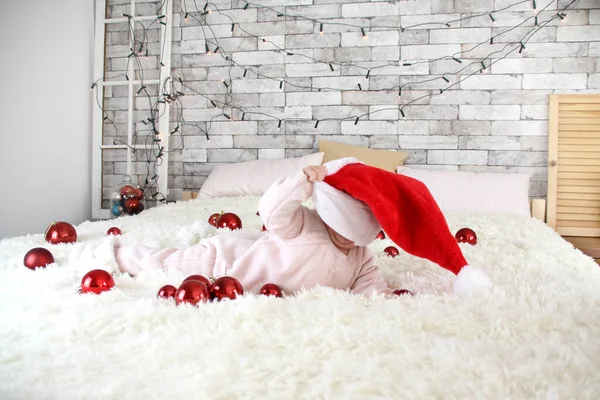 Dziecko w kapeluszu Mikołaja leży na białej puszystej narzutce, a nie łóżku wśród bożonarodzeniowych balów, koncepcji Bożego Narodzenia i Nowego Roku, przytulny dom — Zdjęcie stockowe