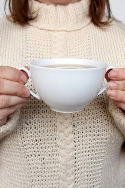 Mujer en un suéter beige de punto cálido sostiene ambas tazas con una taza de té blanco con limón, el concepto de bebidas calientes, acogedor hogar, primer plano — Foto de Stock