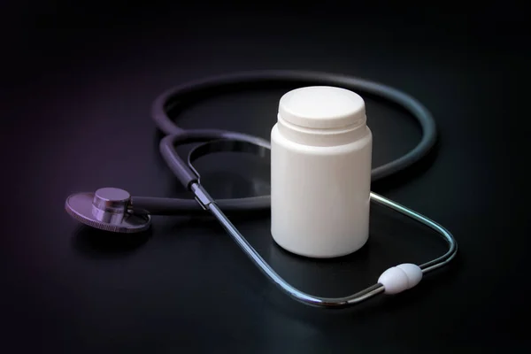 Estetoscópio médico encontra-se em um fundo preto em torno de uma lata de medicina branca vazia, forma para um designer, close-up, espaço de cópia — Fotografia de Stock