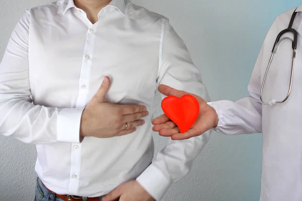 Hombre con una camisa blanca se aferra a su pecho en el área del corazón, a continuación es un médico con un estetoscopio y un modelo de corazón rojo, el concepto de problemas de salud, enfermedades del corazón, cirugía, trasplante de órganos — Foto de Stock