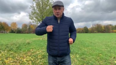 Gri şapkalı ve mavi ceketli bir adam sonbahar parkında spor yapıyor. Eğilimler yapıyor. Yaşlılar için spor konsepti, yakın çekim.