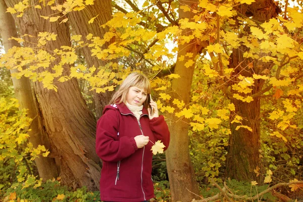 Kobieta ze sceptycznym wyrazem twarzy trzyma telefon w ręku, stoi w jesiennym parku wśród żółtych i pomarańczowych liści klonu, przyciemniane zdjęcie, koncepcja nieprzyjemna rozmowa — Zdjęcie stockowe