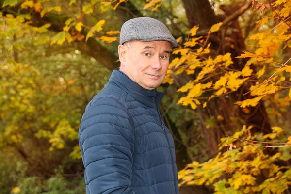 Toned photo of an elderly man in a gray cap and blue jacket, who, with a slight sad smile, stands in an autumn park among yellow and orange leaves of maple trees — Stock Photo, Image