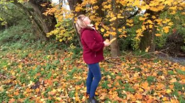 Kot pantolon giymiş orta yaşlı bir kadın ve bordo ceketli. Sonbaharda parkta eğleniyor. Sarı ve portakal ağaçları arasında.