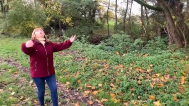 Kot pantolon giymiş orta yaşlı bir kadın ve bordo ceketli. Sonbaharda parkta eğleniyor. Sarı ve portakal ağaçları arasında.