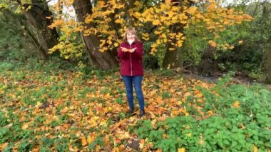 Kot pantolon giymiş orta yaşlı bir kadın ve bordo ceketli. Sonbaharda parkta eğleniyor. Sarı ve portakal ağaçları arasında.