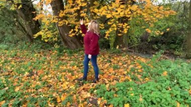 Kot pantolon giymiş orta yaşlı bir kadın ve bordo ceketli. Sonbaharda parkta eğleniyor. Sarı ve portakal ağaçları arasında.