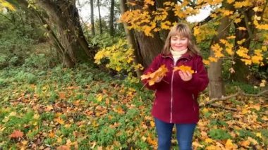 Kot pantolon giymiş orta yaşlı bir kadın ve bordo ceketli. Sonbaharda parkta eğleniyor. Sarı ve portakal ağaçları arasında.