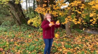 Kot pantolon giymiş orta yaşlı bir kadın ve bordo ceketli. Sonbaharda parkta eğleniyor. Sarı ve portakal ağaçları arasında.