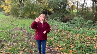 Kot pantolon giymiş orta yaşlı bir kadın ve bordo ceketli. Sonbaharda parkta eğleniyor. Sarı ve portakal ağaçları arasında.