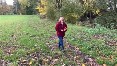 Kot pantolon giymiş orta yaşlı bir kadın ve bordo ceketli. Sonbaharda parkta eğleniyor. Sarı ve portakal ağaçları arasında.