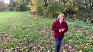 Kot pantolon giymiş orta yaşlı bir kadın ve bordo ceketli. Sonbaharda parkta eğleniyor. Sarı ve portakal ağaçları arasında.