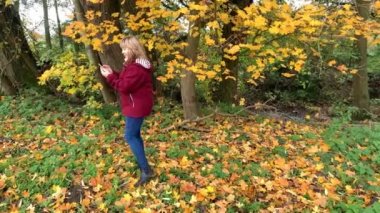 Sonbahar parkında kot pantolon ve bordo ceketli orta yaşlı bir kadın telefonda duygusal olarak konuşuyor, iletişim konsepti, yakın plan.