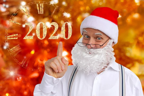 Man in bretels in een pak van de kerstman met een baard, in een hoed toont een vinger op de klok en het jaar 2020, concept Kerstmis, sylvester, op een feestelijke achtergrond — Stockfoto