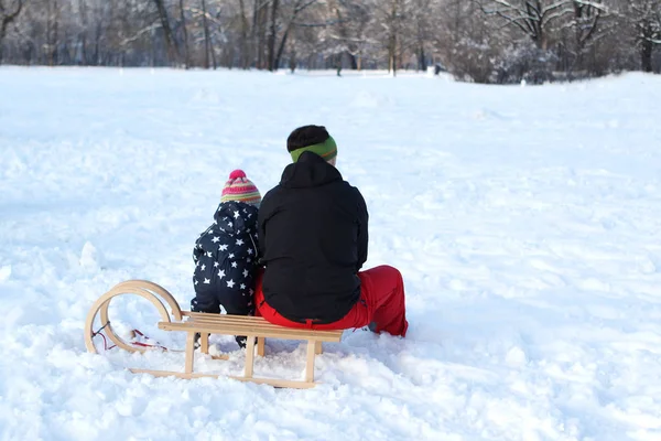 Małe dziecko siedzi z tatą na drewnianych saniach na pierwszym planie pięknego zimowego krajobrazu z białymi śnieżnymi drzewami w parku, głębokimi śladami stóp w śniegu, koncepcją sezonowej aktywności na świeżym powietrzu — Zdjęcie stockowe