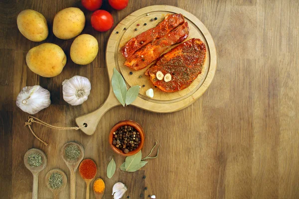 Raw meat, two meat pickled in spices meat medallions on a round wooden board on the table, garlic, bay leaf, potatoes and tomatoes nearby, cooking concept, copy space — Stock Photo, Image