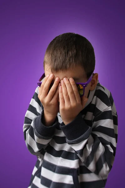 Junge mit dunkler Sonnenbrille und gestreiftem Pullover bedeckt Gesicht mit Angst, lila Hintergrund, Begriff der Bestrafung, psychische und physische Gewalt — Stockfoto