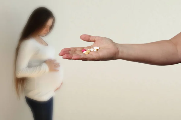 female hand holds out, offering, a handful of colored pills and capsules, close-up, blurred image of a pregnant woman in the background, copy space, concept of pregnancy complications, treatment