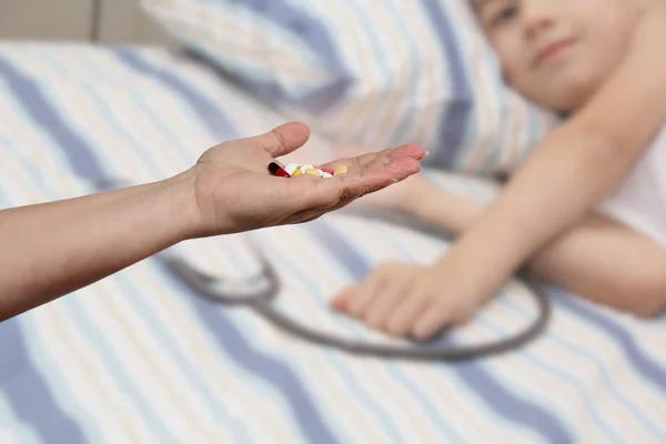 Женская рука протягивает, предлагая, горсть цветных таблеток и капсул, крупным планом, размытое изображение лежащего ребенка на заднем плане, копировальное пространство, концепция медицинского обслуживания, лечение — стоковое фото