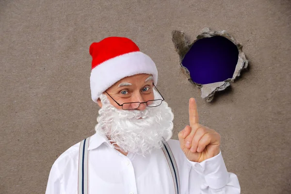 Homem em um traje de Papai Noel com uma barba, em um chapéu, mostra seu dedo através de um buraco rasgado em um papelão marrom vazio, papel artesanal, conceito de sigilo, rastreamento, espionagem, em branco para um designer, close-up — Fotografia de Stock