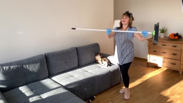 肥胖的中年妇女 围裙中的家庭主妇用拖把进行体育锻炼 用刷子刷打扫房子 运动的概念 体育活动 健康的生活方式 — 图库视频影像