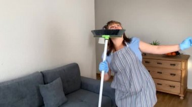 Orta yaşlı bir kadın, önlüklü ve bigudili komik bir ev kadını, paspas ile dans eden, evi temizlemek için bir sopayı fırçalayan, kazanan dansı, ev işi ve ev işi kavramını
