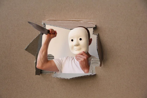 Uomo in maschera bianca sta oscillando un grande coltello, guardando fuori da un buco in cartone, simbolo criminale — Foto Stock