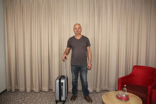 Οριζόντια φωτογραφία ενός άνδρα, τουρίστα, στέκεται με ένα χαμόγελο σε ένα δωμάτιο ξενοδοχείου με μια βαλίτσα από το παράθυρο, τουριστική έννοια — Φωτογραφία Αρχείου