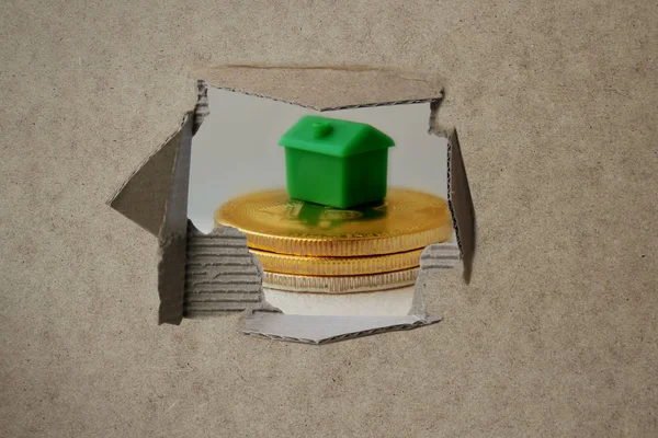 Through a torn hole in an empty brown cardboard blank a stack of metal coins is visible on which stands a green mock-up of a house, a concept of business and buying real estate, close-up — 스톡 사진