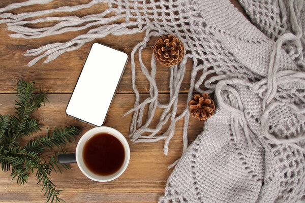 flat lay background with a mug of tea, white smart phone with blank screen, shawl with fringe, coniferous branches and dried orange cinnamon wood, concept winter mood, winter is love, copy space
