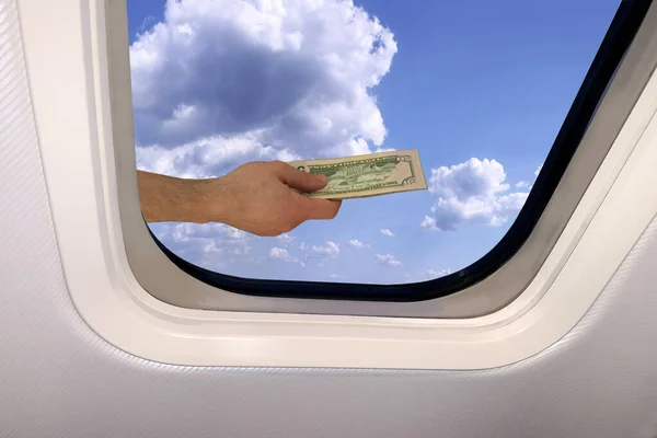 Mano masculina con un paquete de billetes de dólares en el fondo de un hermoso paisaje celestial de cielo azul con nubes esponjosas detrás del ojo de buey de un avión, concepto de viaje — Foto de Stock