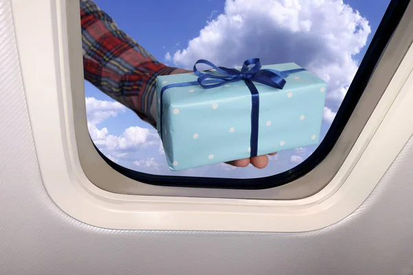 Мужская рука с подарком в коробке на фоне прекрасного небесного ландшафта голубого неба с пушистыми облаками за иллюминатором самолета, концепцией подарков и дорожных бонусов — стоковое фото