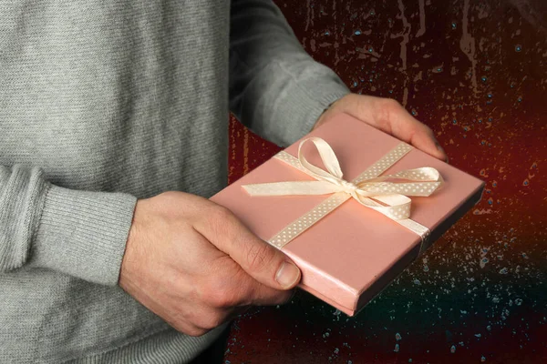 Mężczyzna trzyma prezent w ręku w różowym pudełku z satynową wstążką na ciemnym tle, koncepcja wakacje — Zdjęcie stockowe