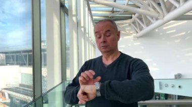 Havaalanı lobisinin parlak binasında sırt çantalı bir turist pencerenin yanında duruyor ve sokağa bakıyor, saatine bakıyor. Bekleme ve uçuş gecikmeleri kavramı.